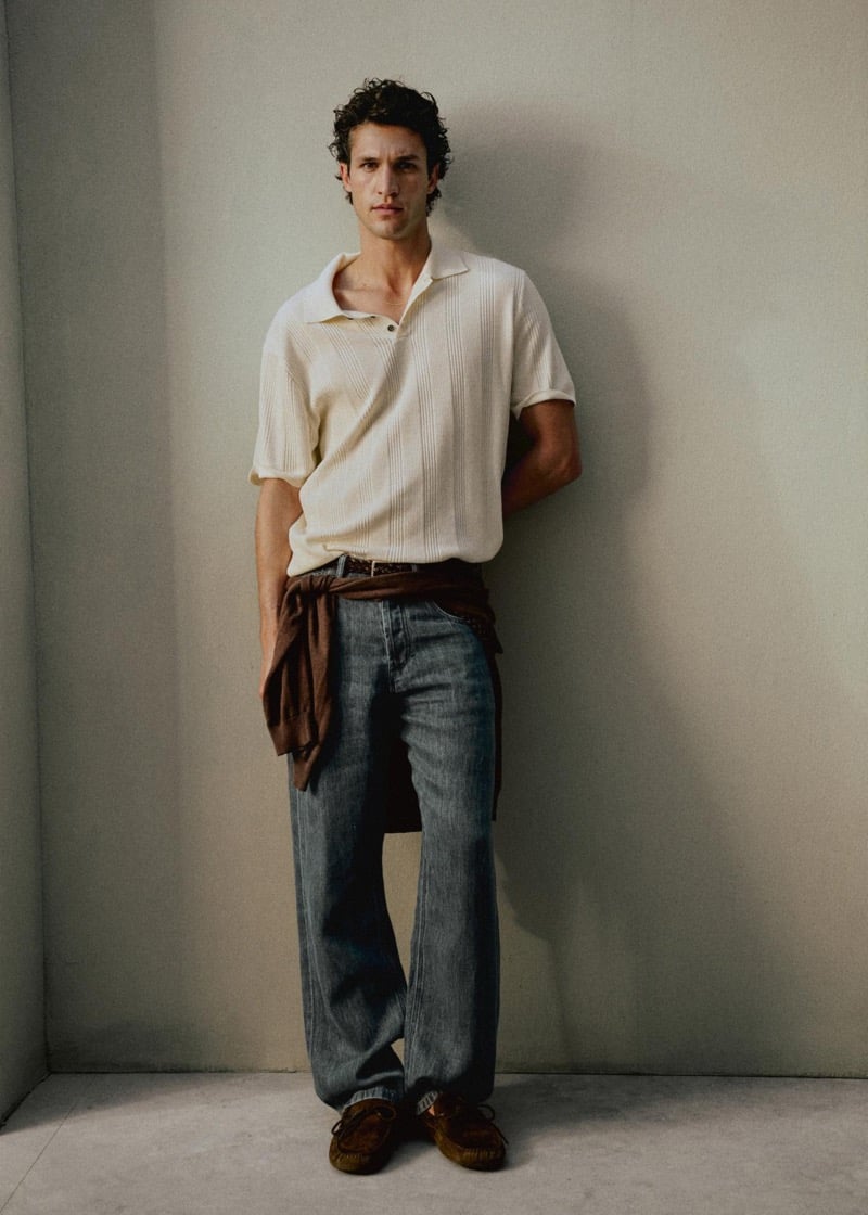 Francisco Henriques stands against a sand-toned wall in a ribbed cream knit polo with a brown sweater tied around his waist, baggy jeans, a braided leather belt, and brown suede loafers