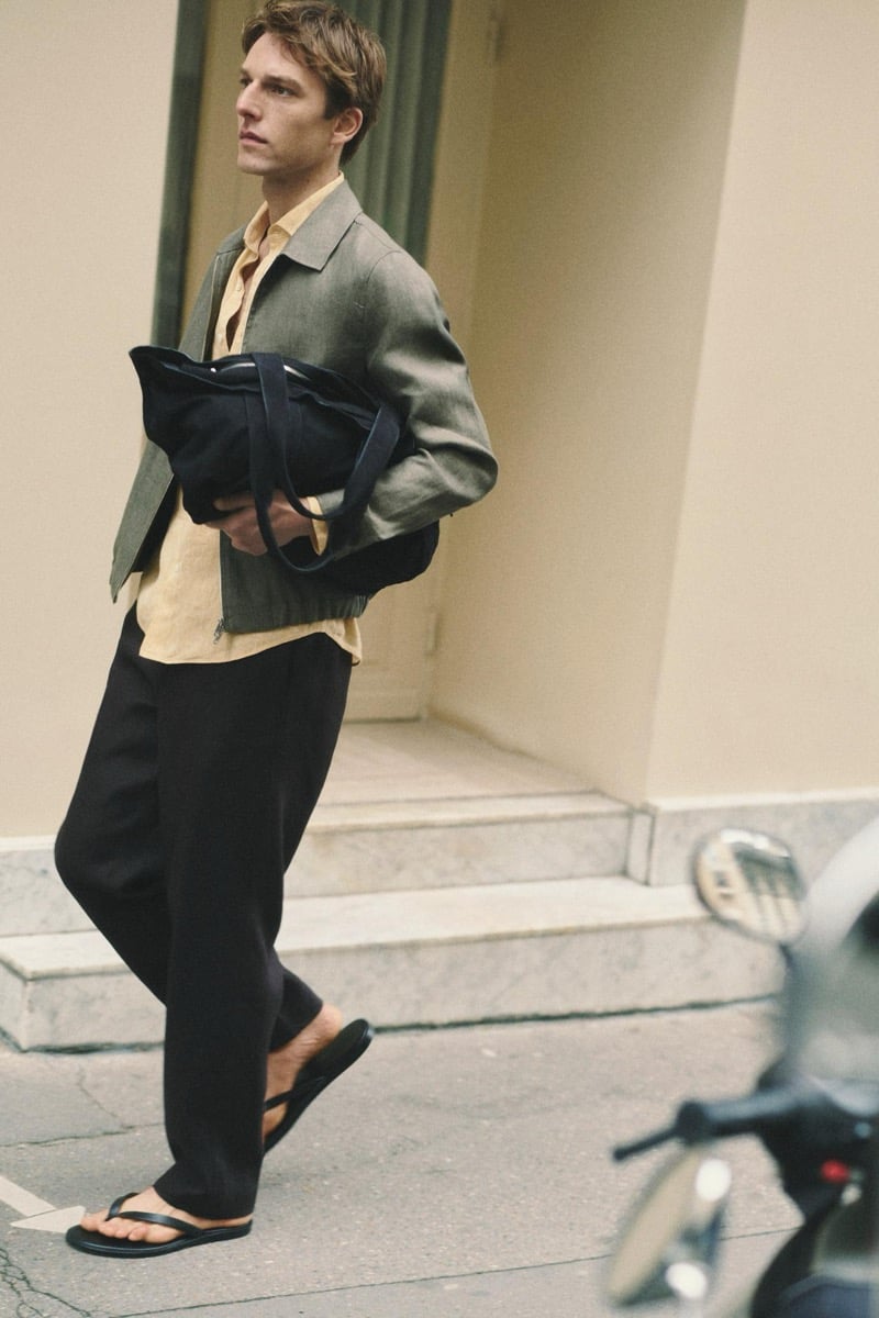 Quentin Demeester walking a Paris sidewalk in a Massimo Dutti linen blouson and pleated trousers, a soft tote tucked under one arm and leather flip-flops on his feet