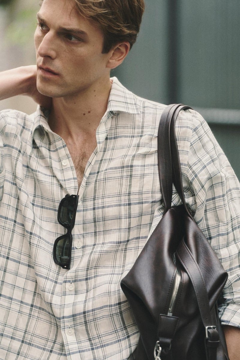 Quentin Demeester in a checked Massimo Dutti linen shirt with rectangular sunglasses hooked at the placket and a leather tote on his shoulder