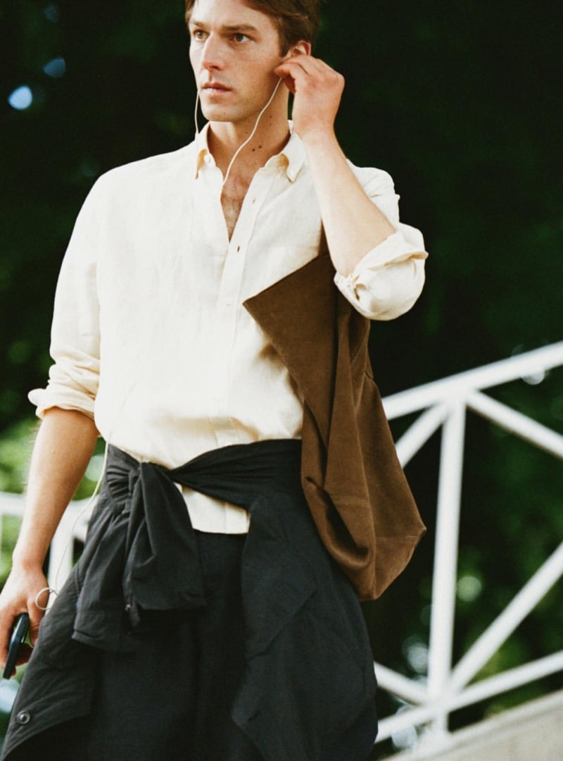 Quentin Demeester walking with wired earbuds in a Massimo Dutti linen shirt, a suede tote on the shoulder and an overshirt knotted at the waist