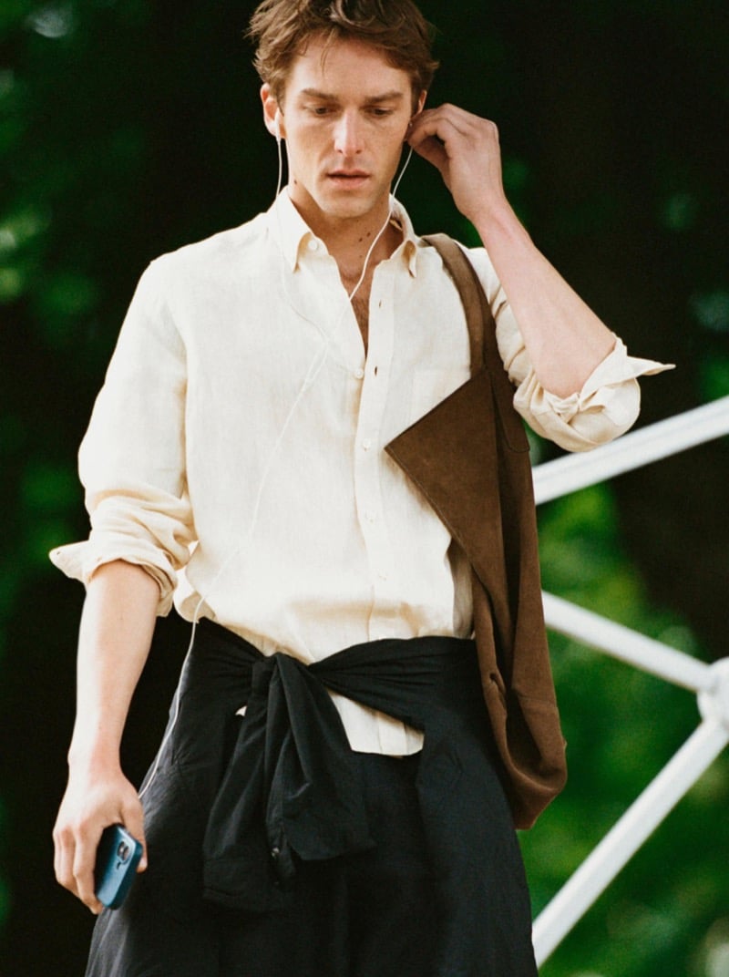 Quentin Demeester walking with wired earbuds in a Massimo Dutti linen shirt, a suede tote on the shoulder and a lightweight overshirt knotted at the waist