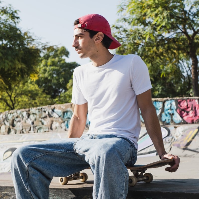Young man in a red ball cap and a clean white crewneck tee sitting on a graffiti-walled skatepark ledge with his skateboard, the kind of staple tee built for daily rotation
