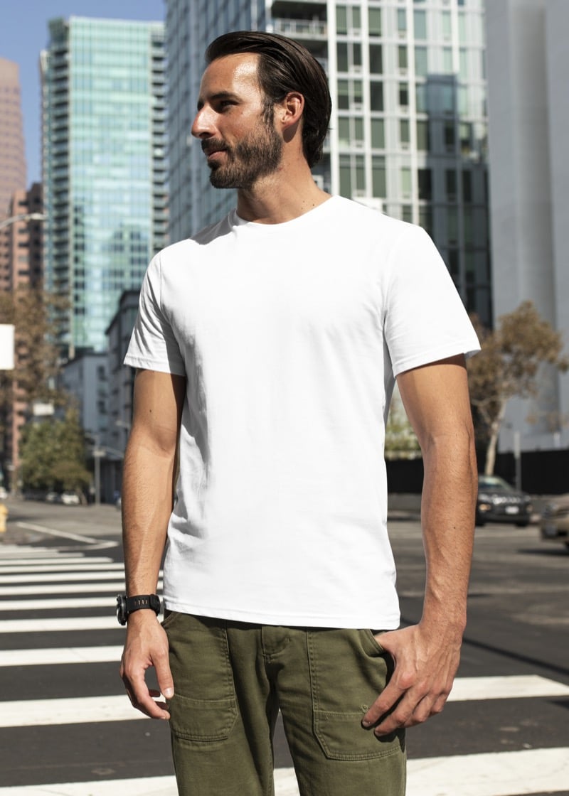 Bearded man in a fitted white crewneck T-shirt and olive cargo pants standing on a city crosswalk, the shoulder seam landing exactly at the edge of the shoulder