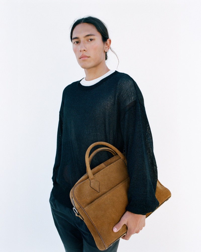 Cherokee Jack in a sheer black open-weave linen knit layered over a white t-shirt, carrying the Métier Closer Slim briefcase in tobacco Marrakech suede tucked against his ribs