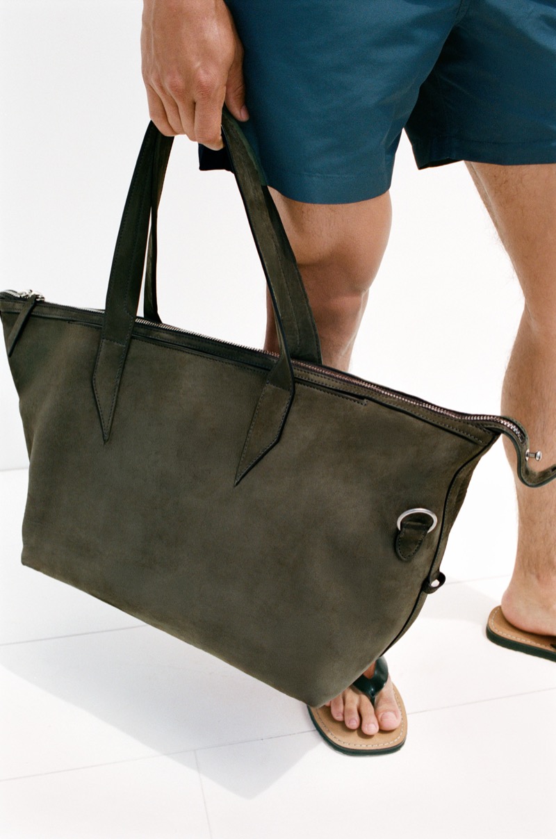 Cropped view of a man in teal swim shorts and leather thong sandals on a white tiled floor, holding the Métier Nomad All Day tote in deep olive Corfu suede at his side