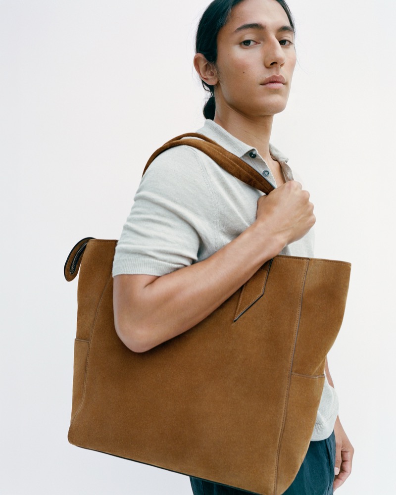 Cherokee Jack standing against a soft white wall in a heather gray short-sleeve polo and dark trousers, his hair pulled back, with the Métier Passerby North-South tote in tobacco Marrakech suede resting against his right shoulder