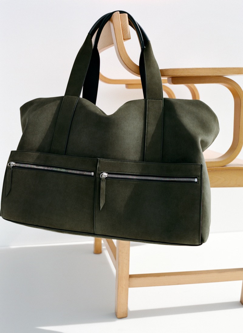 The Métier Racer 53 weekender in deep olive Corfu suede, resting across the curved arms of a blonde bentwood lounge chair, with twin front zip pockets visible