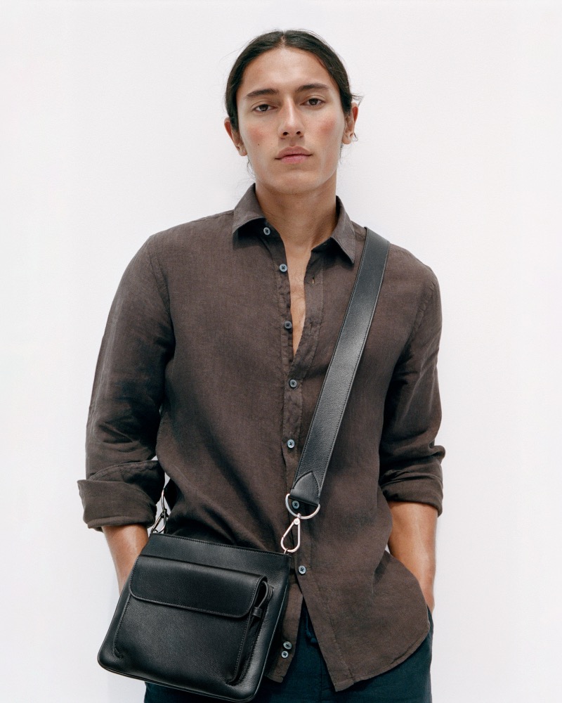 Cherokee Jack in a chocolate brown linen shirt and dark trousers against a white wall, the Métier Slide Messenger 21 in buffalo black grained leather worn crossbody at his hip