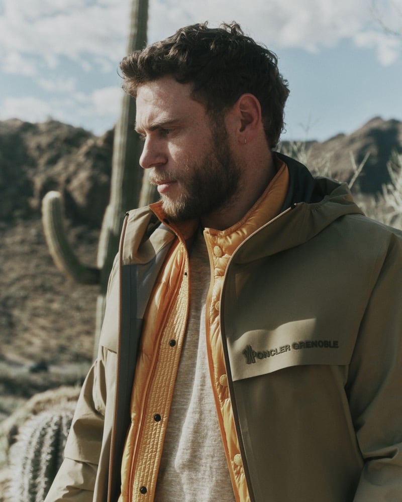 Gus Kenworthy in profile against the Tucson Mountain Park desert with saguaro cacti behind him, wearing a sand hooded shell jacket with a Moncler Grenoble chest pocket, layered over a marigold quilted puffer vest and an oatmeal crewneck tee