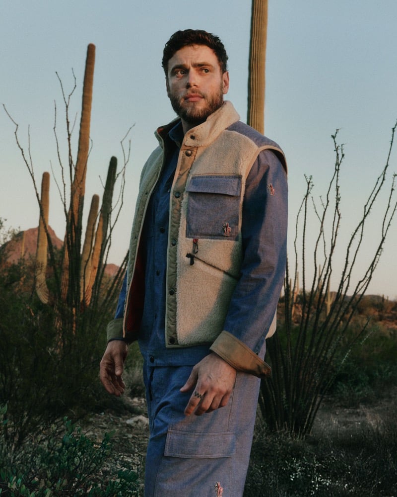 Gus Kenworthy standing among saguaro cacti and ocotillo branches at dusk, in a cream teddy-panel utility vest with chambray-trim pockets worn over a matching chambray work shirt and chambray cargo trousers, the Moncler Grenoble pin badge stitched at the chest