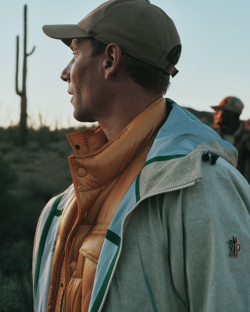 Richard Permin seen in profile in a baseball cap, layered in a color-block shell jacket with a Moncler Grenoble logo on the sleeve, over a marigold quilted puffer vest in the desert at golden hour