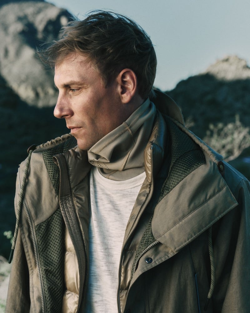 Richard Permin in a tight crop against the rocky outcrops of Tucson Mountain Park, wearing an olive-green technical field jacket with mesh chest panels, layered over a champagne quilted puffer vest, a marled cream crewneck tee, and a sand technical balaclava neck gaiter