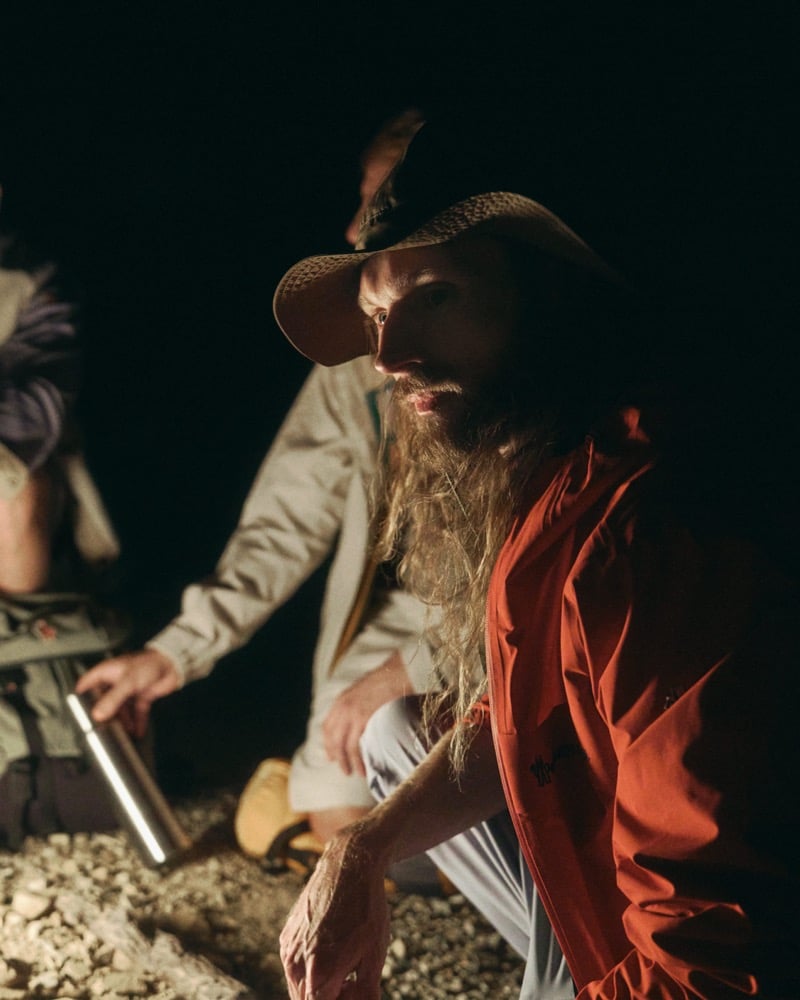 Aiden Andrews crouched at a campfire after dark in a straw cowboy hat, wearing a brick-red lightweight zip-up windbreaker with the Moncler Grenoble script embroidered at the chest and cream technical trousers, holding a stainless steel canister beside another camper in a cream jacket