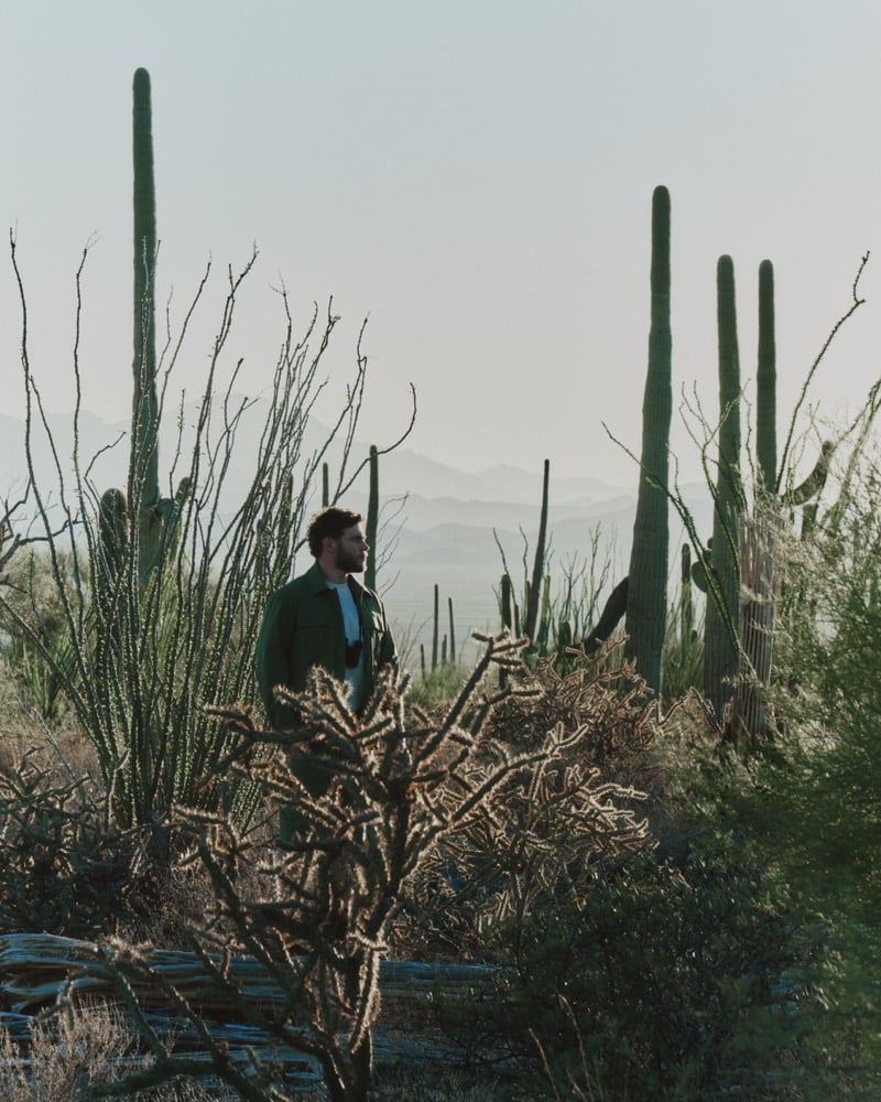 Gus Kenworthy walking through a cluster of saguaro and cholla cacti with the Tucson Mountains in the distance, wearing dark sunglasses and a forest-green chest-pocket field shacket over a pale blue tee, the desert plants framing him in the foreground
