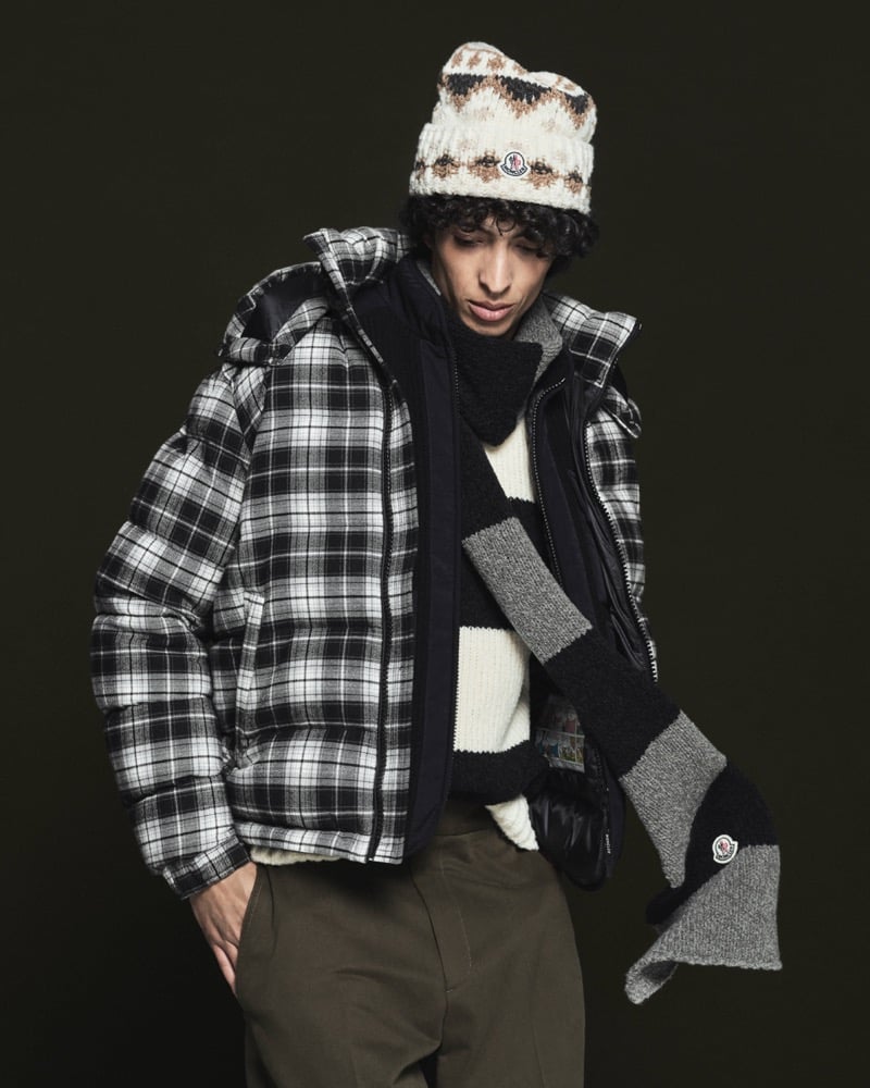 Model Nour Boudaakat in a cream Fair Isle knit beanie with Moncler logo wears a black and white tartan padded jacket with navy nylon panels, a white and black striped knit sweater, gray and black striped wool scarf, black quilted glove, and olive trousers
