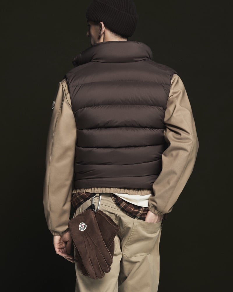 Back view of a model in a black ribbed beanie wearing a chocolate brown quilted down vest over a camel cotton harrington jacket, plaid shirt, and khaki trousers, with brown suede gloves clipped on a carabiner