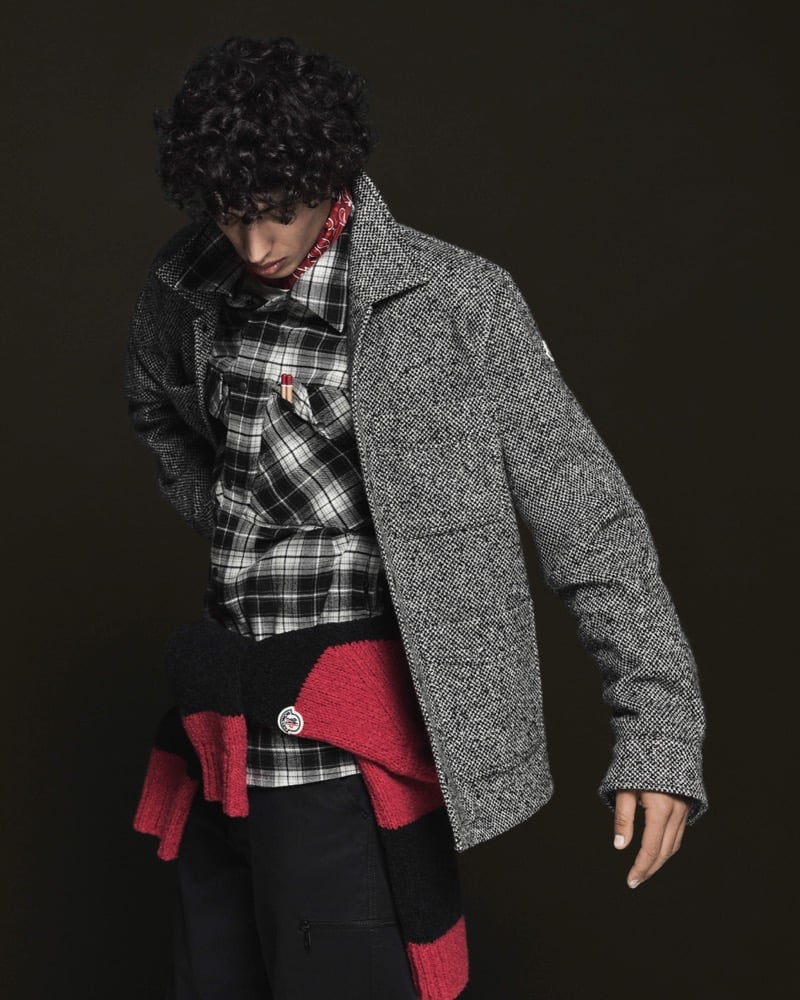 Model Nour Boudaakat in a black and white salt and pepper tweed jacket with patch pockets over a black and white plaid flannel shirt, a red and black striped knit sweater tied at the waist, a red paisley bandana, and black cargo trousers