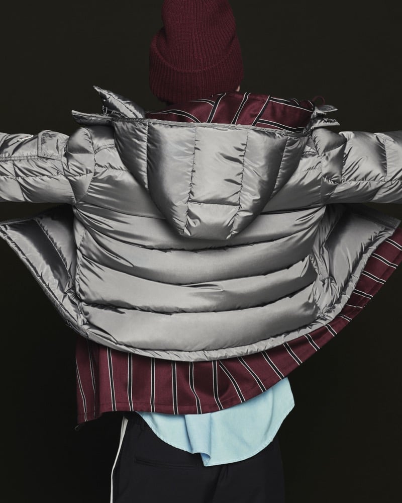 Back view of a model in a burgundy ribbed beanie wearing an open silver-gray down puffer jacket over a burgundy striped shirt, pale blue button-down, and black trousers