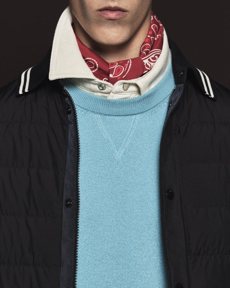 Close-up of a black quilted nylon overshirt tipped in black and white stripes, layered over a sky blue knit crewneck, cream button shirt, and red paisley bandana