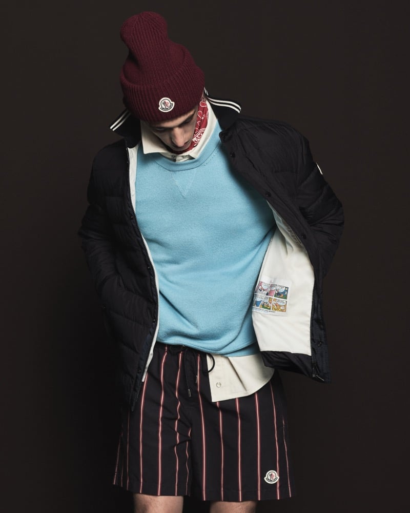 Model in a burgundy ribbed beanie with Moncler logo wears a black quilted down jacket with a white lining and comic-strip patch, sky blue crewneck sweater, cream button shirt, red paisley bandana, and navy drawstring shorts with red stripes
