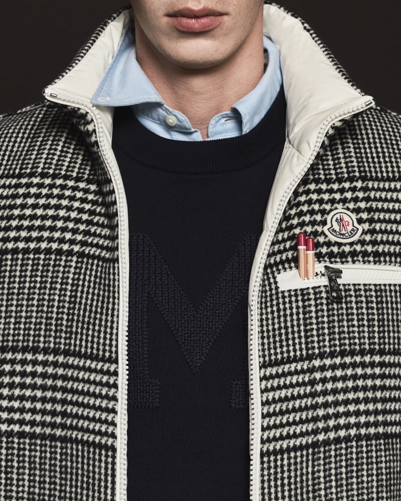Close-up of a black and cream Prince of Wales check zip jacket with cream nylon trim and pencils tucked in the chest pocket, layered over a navy crewneck sweater with a tonal M letter motif and a pale blue oxford shirt