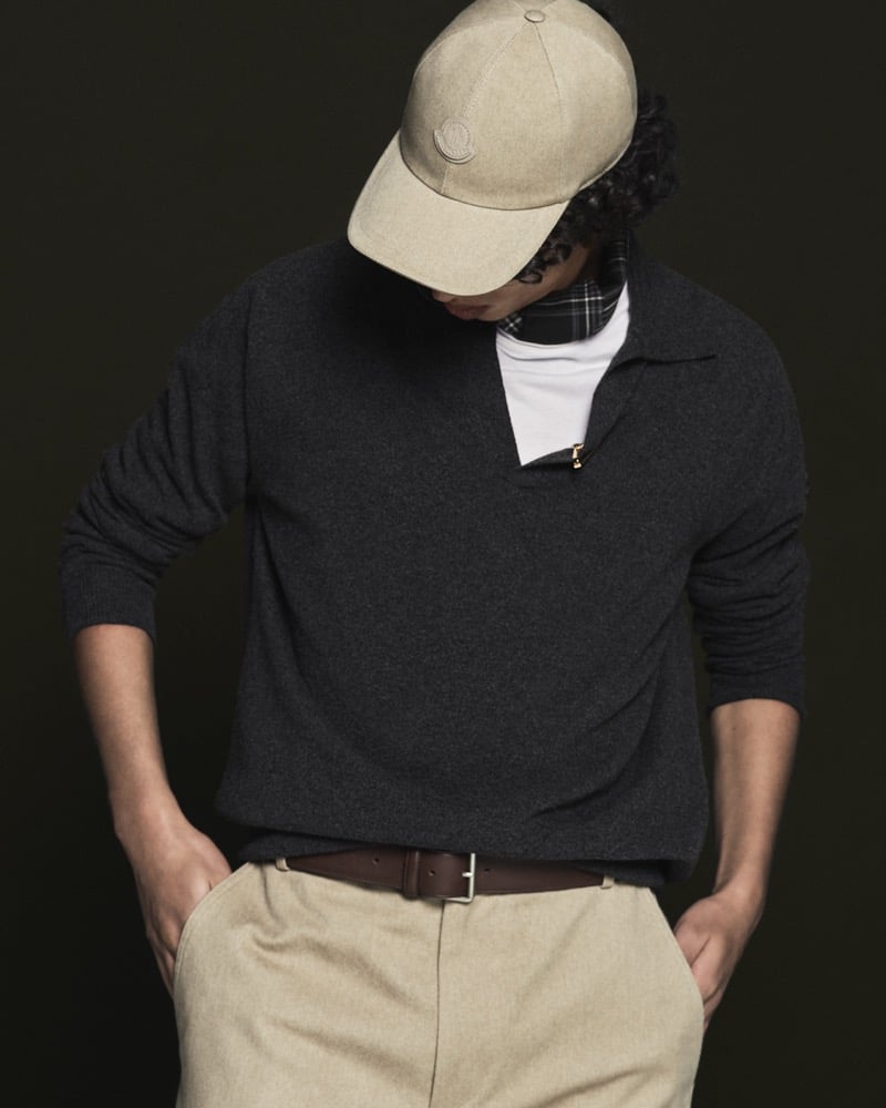 Model Nour Boudaakat in a sand canvas baseball cap with Moncler logo wears a charcoal half-zip cashmere sweater with a gold zipper pull over a white tee, paired with a brown leather belt and oatmeal flannel trousers