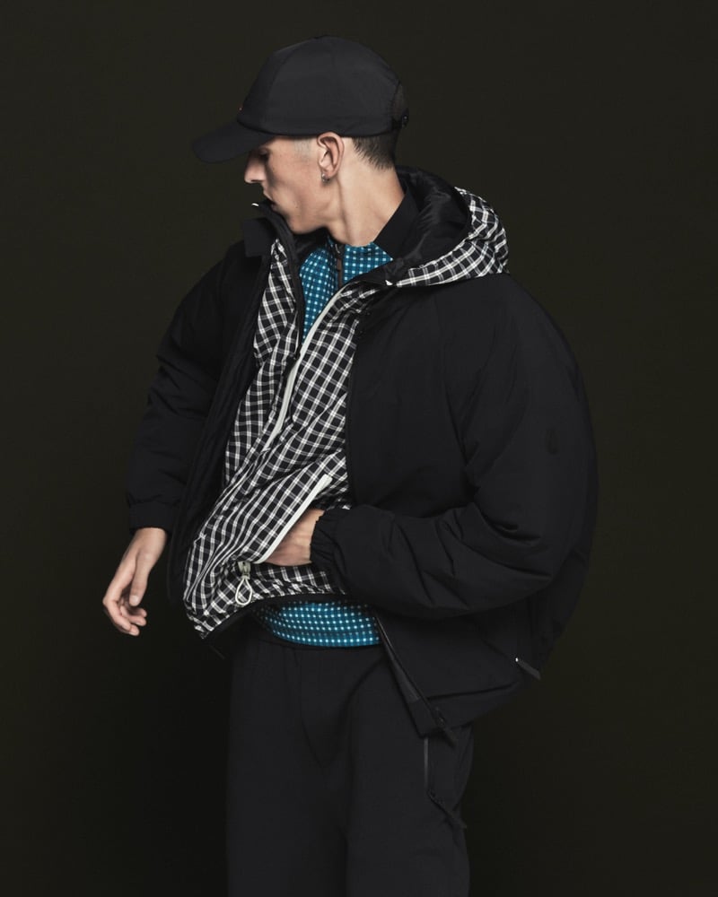 Model in a black cap wears an oversized black nylon down jacket over a black and white gingham zip jacket, teal blue grid-pattern knit, and black technical trousers