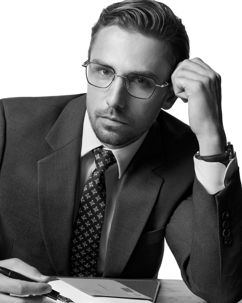 James Yates wears slim rectangular metal optical frames with a tailored two-button suit and a patterned medallion tie, seated at a desk holding a Montblanc pen over an open notebook in a black-and-white portrait