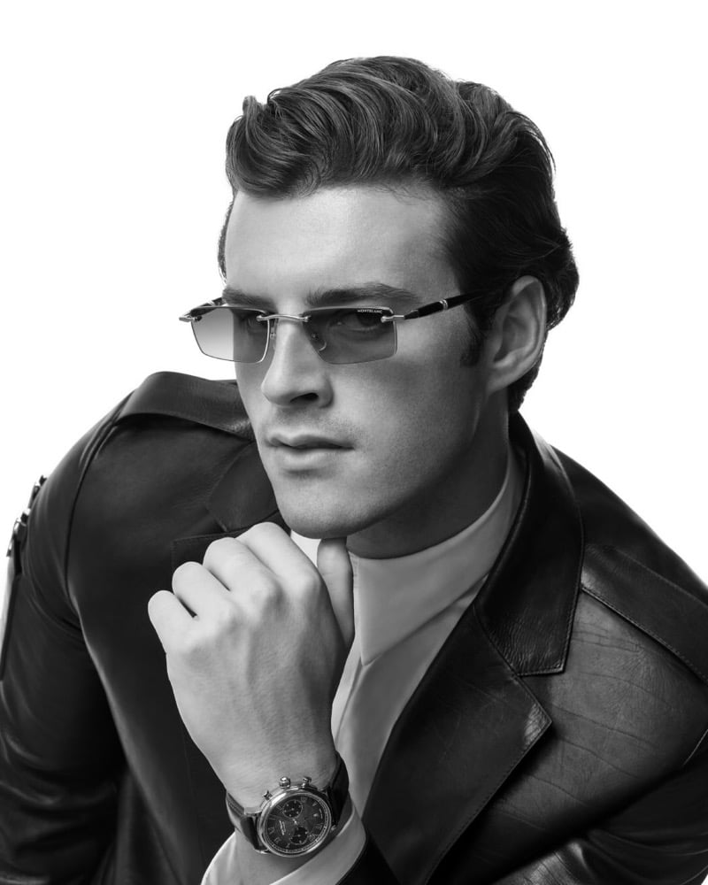 Aiden Stonehouse wears rimless tinted sunglasses and a leather jacket, his hand near his chin revealing a Montblanc chronograph watch on his wrist in a black-and-white portrait