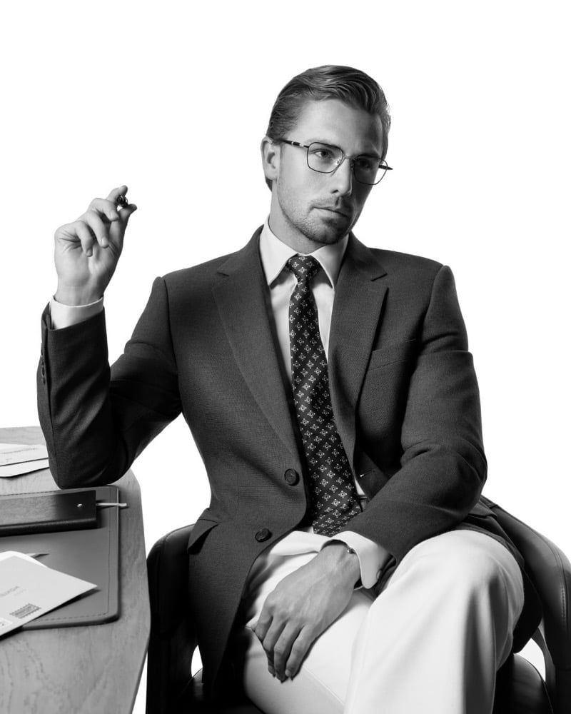 James Yates sits in a tailored two-button suit with a patterned tie and rectangular metal optical frames, holding a pen mid-air beside a leather notebook in a black-and-white portrait
