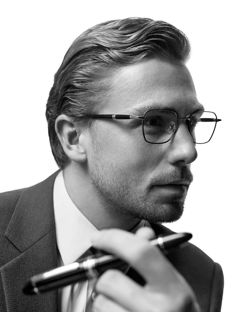 Close-up profile of James Yates in square metal optical frames with engraved temple detailing, holding a signature Montblanc rollerball pen near his chest in a black-and-white portrait