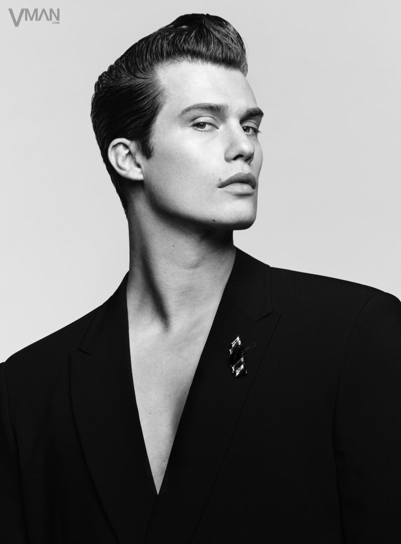 Nicholas Galitzine looks past the camera with slicked-back hair in a sharply tailored black Giorgio Armani suit jacket with a jeweled brooch on the lapel
