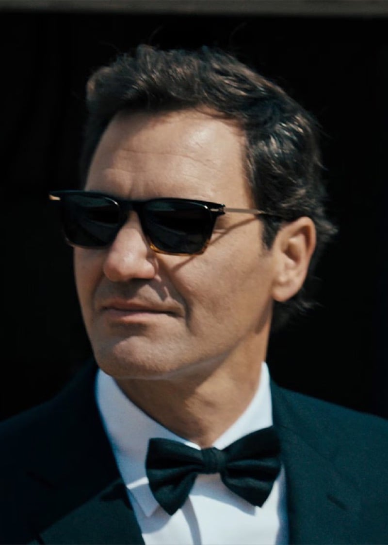 Close portrait of Roger Federer in a black tuxedo jacket, crisp white dress shirt, and black silk bow tie, wearing rectangular Oliver Peoples sunglasses with a tortoise-shell lower rim and slim metal temples