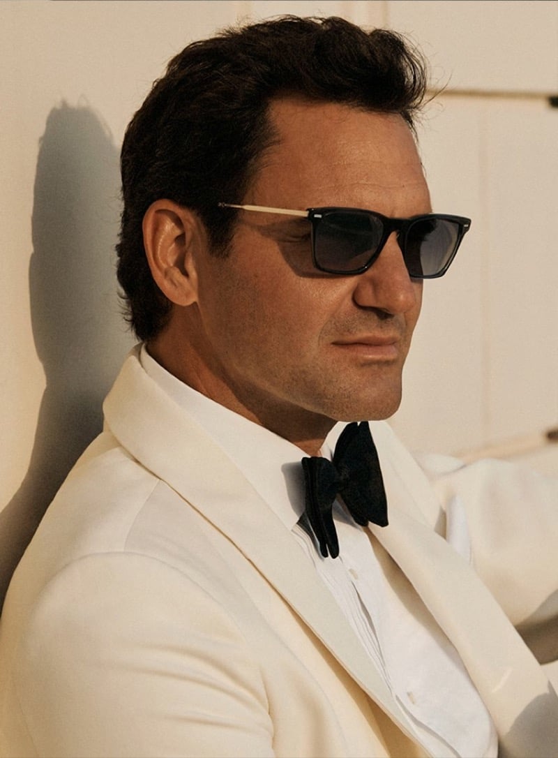 Side profile of Roger Federer against a sunlit cream wall in a white shawl-lapel dinner jacket, white shirt, and black silk bow tie, wearing squared Oliver Peoples sunglasses with thin gold metal temples and an upswept lens line