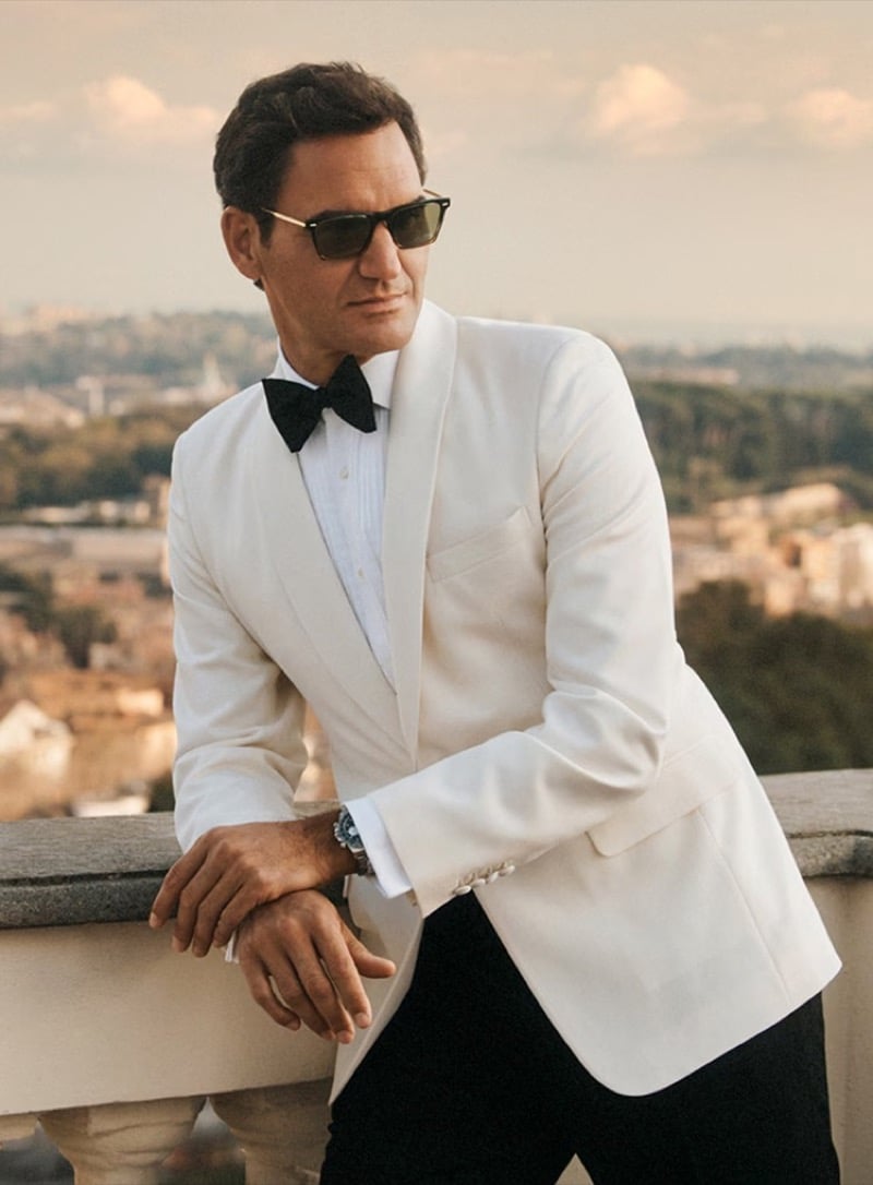 Roger Federer leaning against a stone balustrade with the Roman skyline behind him, dressed in a white dinner jacket, black bow tie, white pleated shirt, and black trousers, wearing angular double-bridge Oliver Peoples sunglasses with metal detailing