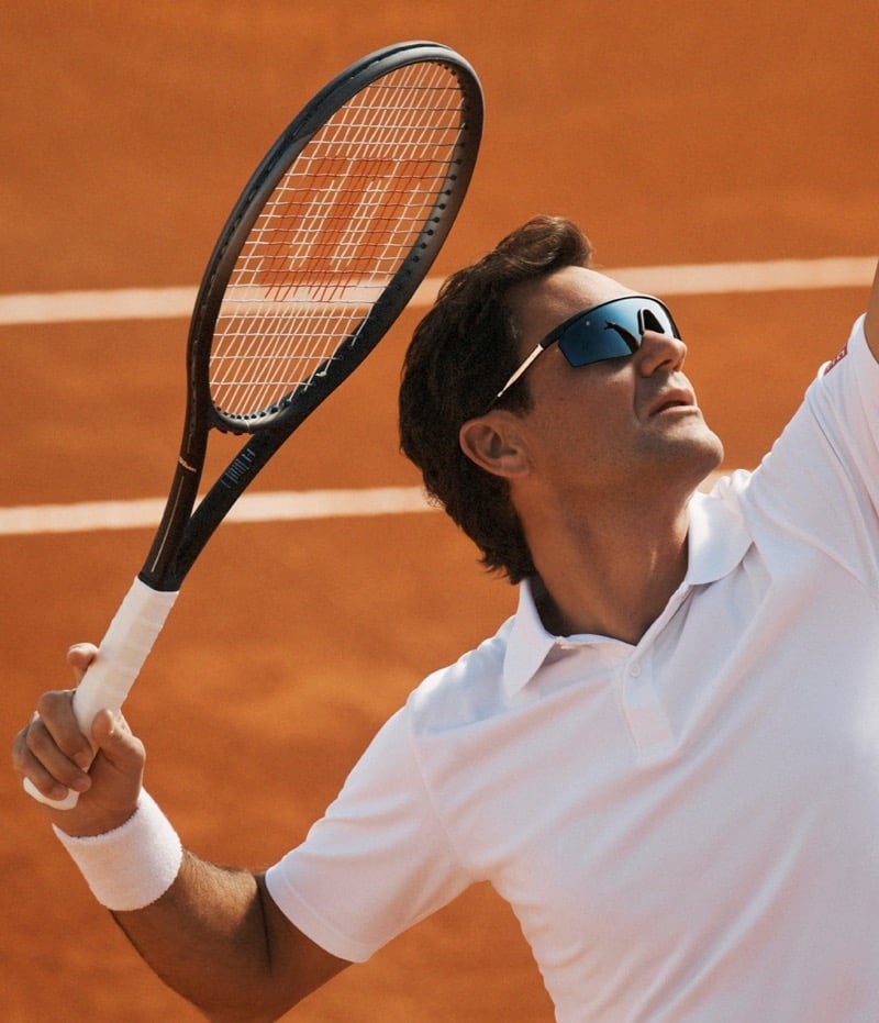 Roger Federer mid-serve on red clay in a white tennis polo and matching wristband, racquet raised overhead, wearing wrapped shield-style Oliver Peoples sunglasses with mirrored color-enhancing lenses
