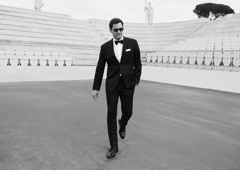 Black and white image of Roger Federer walking through the Stadio dei Marmi at the Foro Italico in a black tuxedo and bow tie, wearing rectangular Oliver Peoples sunglasses