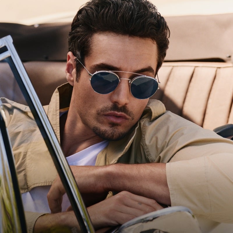 Levi Dylan reclines against the tan leather seat of a vintage convertible in a tan field jacket, wearing round silver-framed sunglasses with blue lenses