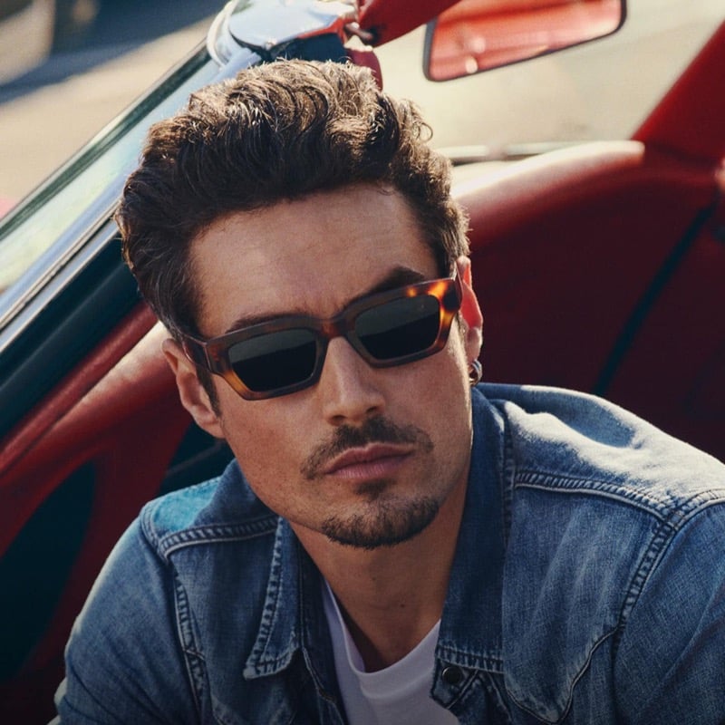Levi Dylan leans against a red vintage convertible in a denim trucker jacket, wearing rectangular tortoise-frame sunglasses