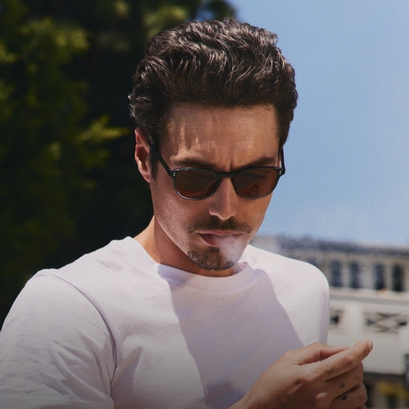 Levi Dylan looks down at his hand in a white tee against a blue sky, wearing dark gradient-lens rectangular sunglasses