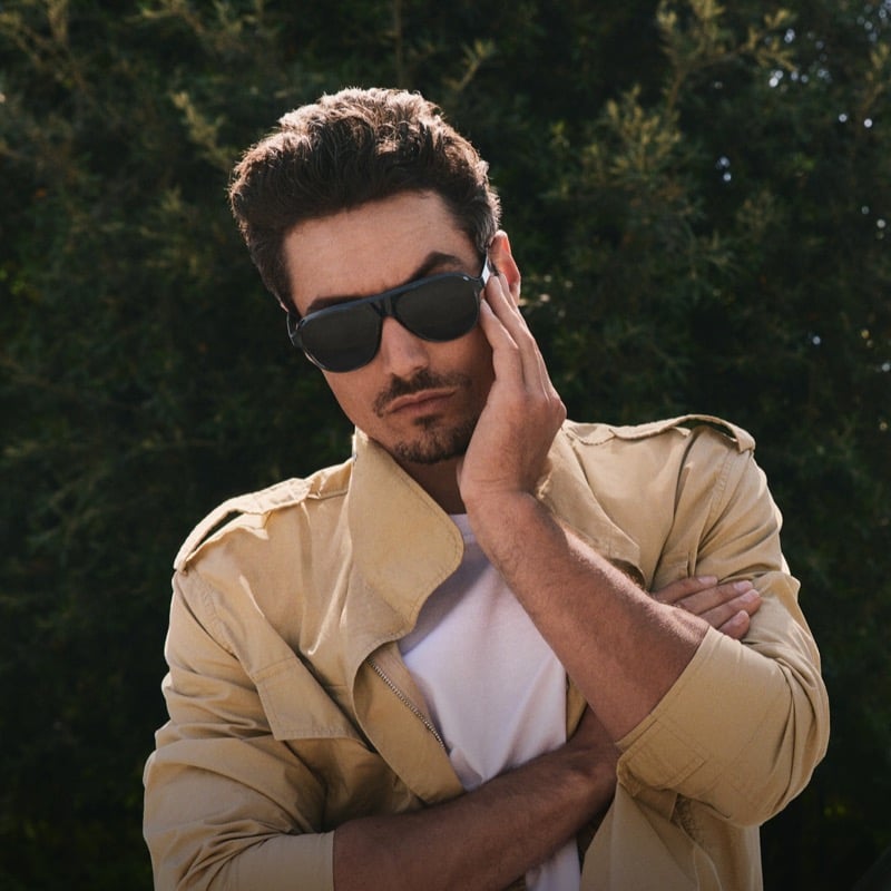 Levi Dylan stands with arms folded in a tan field jacket over a white tee, wearing dark sunglasses in front of a tall hedge