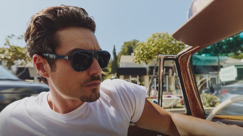 Levi Dylan rests an arm on the door of a vintage convertible in a white tee, wearing Oliver Peoples Paul Newman sunglasses with a Sunset Plaza sign in the distance