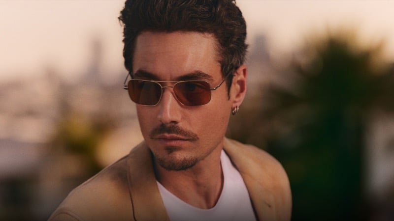 Close-up of Levi Dylan at dusk in a tan jacket over a white tee, wearing Oliver Peoples Victory II sunglasses with gold metal frames and cognac-tinted lenses