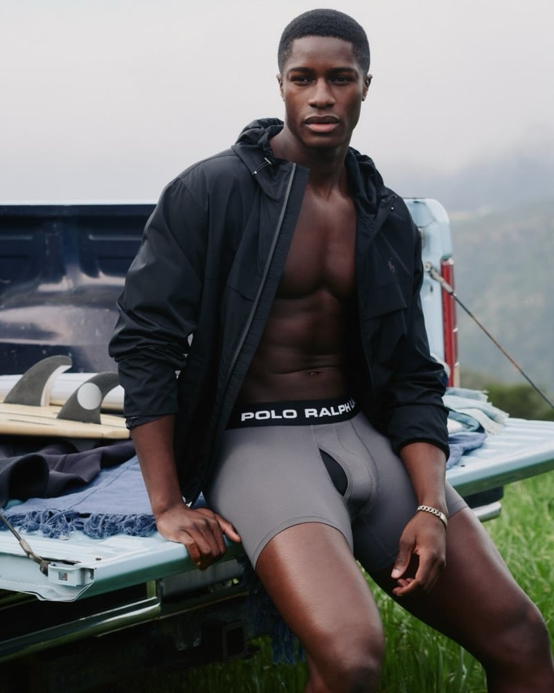 Model Mukasa Kakonge in a black hooded windbreaker and gray Polo Ralph Lauren logo-waistband boxer briefs, seated on the tailgate of a vintage pickup truck with a surfboard beside him