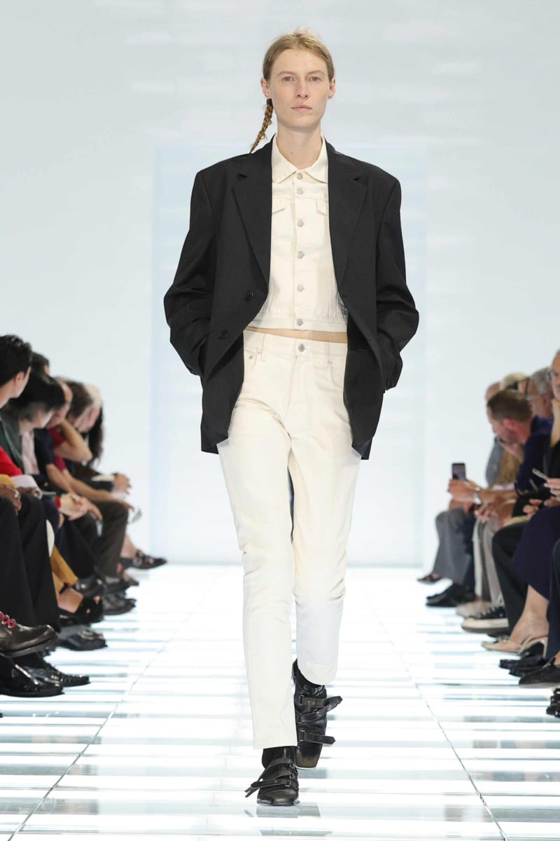 A Prada model walks the spring 2027 runway in an oversized black wool blazer over a cropped ivory denim jacket and matching ivory jeans, finished with black multi-strap leather shoes