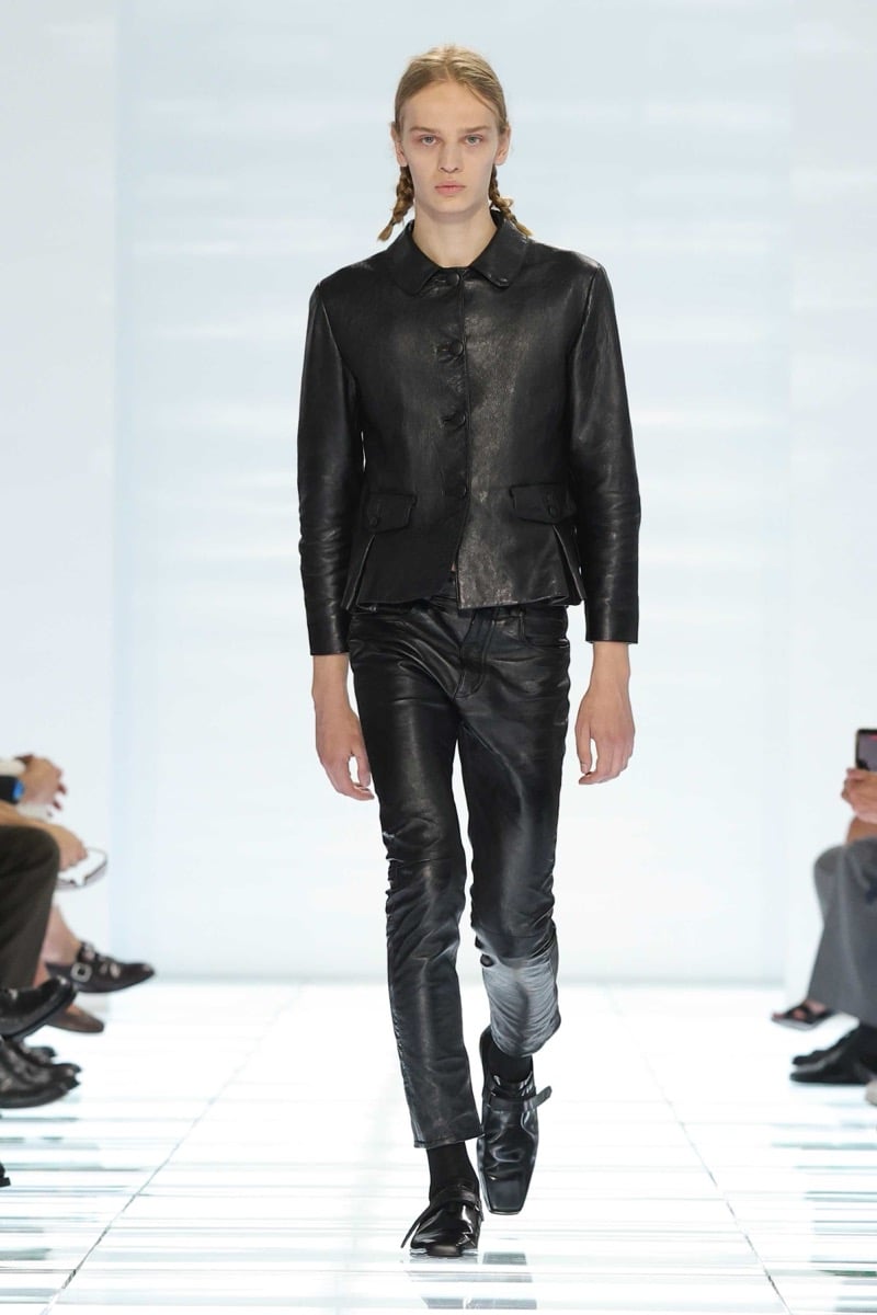 A Prada model walks the spring 2027 runway in a fitted black leather jacket with peter pan collar and flap pockets over slim black leather trousers and black multi-strap leather shoes