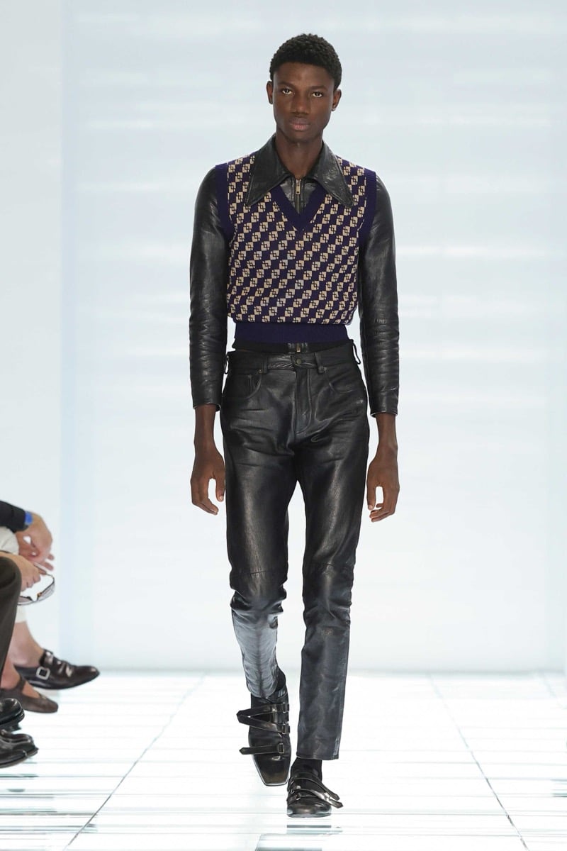 A Prada model walks the spring 2027 runway in a navy and tan diamond geometric knit vest layered over a black leather collared shirt, paired with slim black leather trousers and black strap shoes
