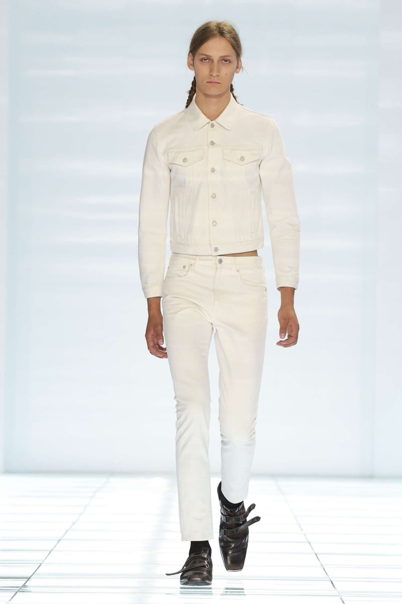 A Prada model walks the spring 2027 runway in a cropped ivory denim jacket and matching ivory slim jeans, finished with black multi-strap leather shoes