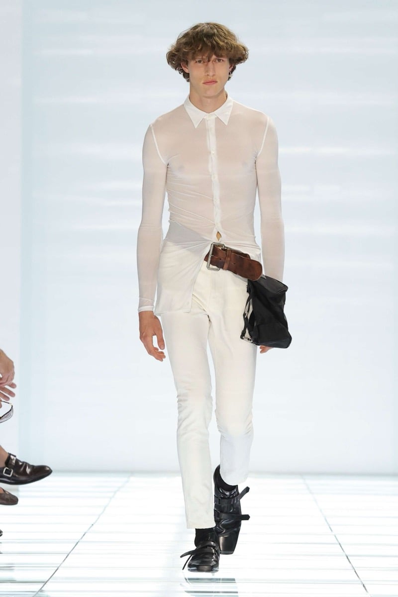 A Prada model walks the spring 2027 runway in a sheer white long-sleeve button-up shirt knotted at the waist over a thick brown leather belt, paired with slim white trousers, a black leather pouch, and black strap shoes