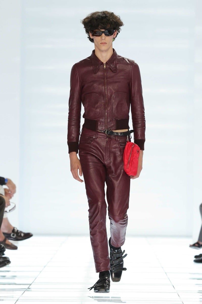 A Prada model walks the spring 2027 runway in narrow black sunglasses, a cropped burgundy leather bomber jacket with knit cuffs, and matching burgundy leather trousers, carrying a red Prada nylon pouch on the belt